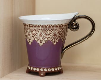 May include: A purple ceramic mug with a white lace design and a silver handle. The mug has a scalloped rim and sits on three small feet.