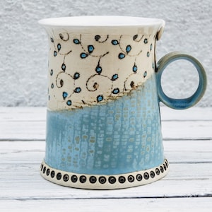 Puede incluir: Una taza de cerámica hecha a mano con un diseño floral azul y blanco. La taza tiene un esmalte azul texturizado y un borde blanco. El asa es azul.