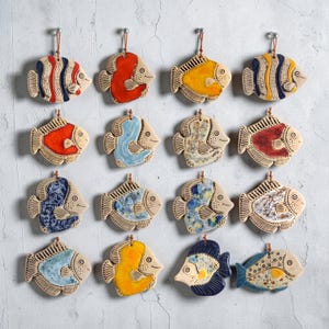 May include: A collection of ceramic fish ornaments in various colors, including red, blue, yellow, and white. Each fish has a unique design and is hung by a brown string. The ornaments are displayed against a light gray background.