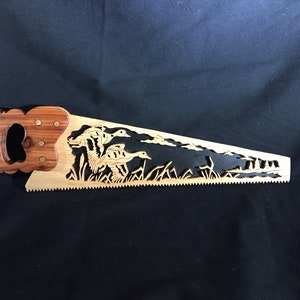 May include: Wooden hand saw with a decorative duck scene cut into the blade. The handle is a darker wood, and the blade is a lighter wood. The saw teeth are visible along the bottom edge. The ducks are in a grassy area.
