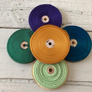 May include: A collection of colorful spools of ribbon in shades of green, gold, teal, purple, and light green. Each spool is neatly wound and has a wooden core. The ribbons are arranged on a light-colored wooden surface.