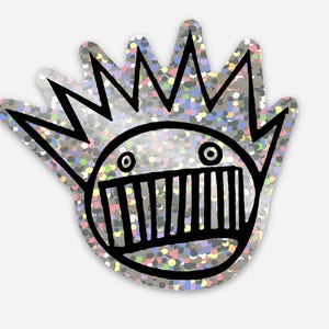 May include: A silver sticker featuring a cartoon face design. The face has a wide, toothy grin, two round eyes, and a crown-like shape above the head. The sticker has a glittery, multi-coloured background.