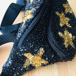 Op de afbeelding: Een zwarte heuptas met een gouden paillettenontwerp. De tas heeft een ritssluiting en een verstelbare riem.