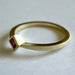 May include: A simple gold band ring with a small square-shaped ruby gemstone set in the band.