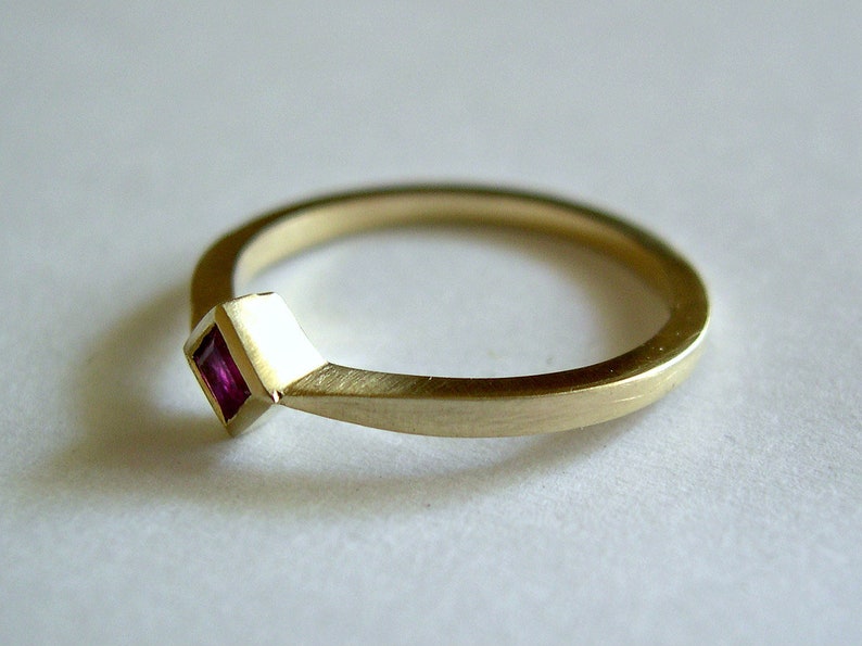 May include: A simple gold band ring with a small square-shaped ruby gemstone set in the band.