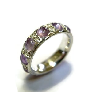 May include: A silver band ring with a row of purple gemstones set in the band. The ring has a decorative design around the gemstones.