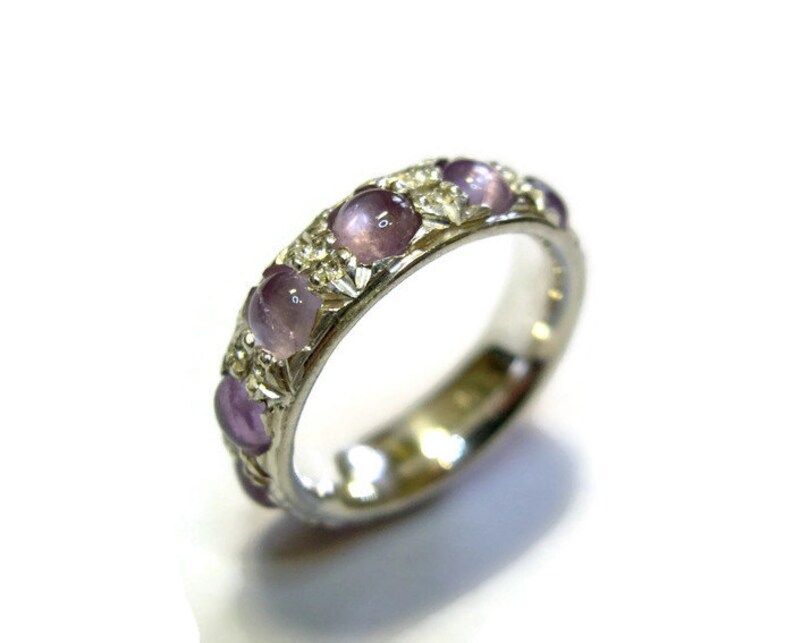 May include: A silver band ring with a row of purple gemstones set in the band. The ring has a decorative design around the gemstones.