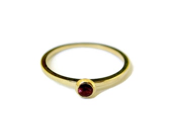 May include: A simple gold band ring with a single, round, dark red gemstone.