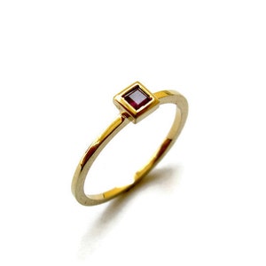May include: A gold ring with a small, square, red gemstone set in a gold frame.