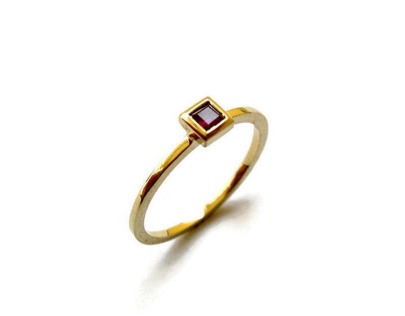 May include: A gold ring with a small, square, red gemstone set in a gold frame.