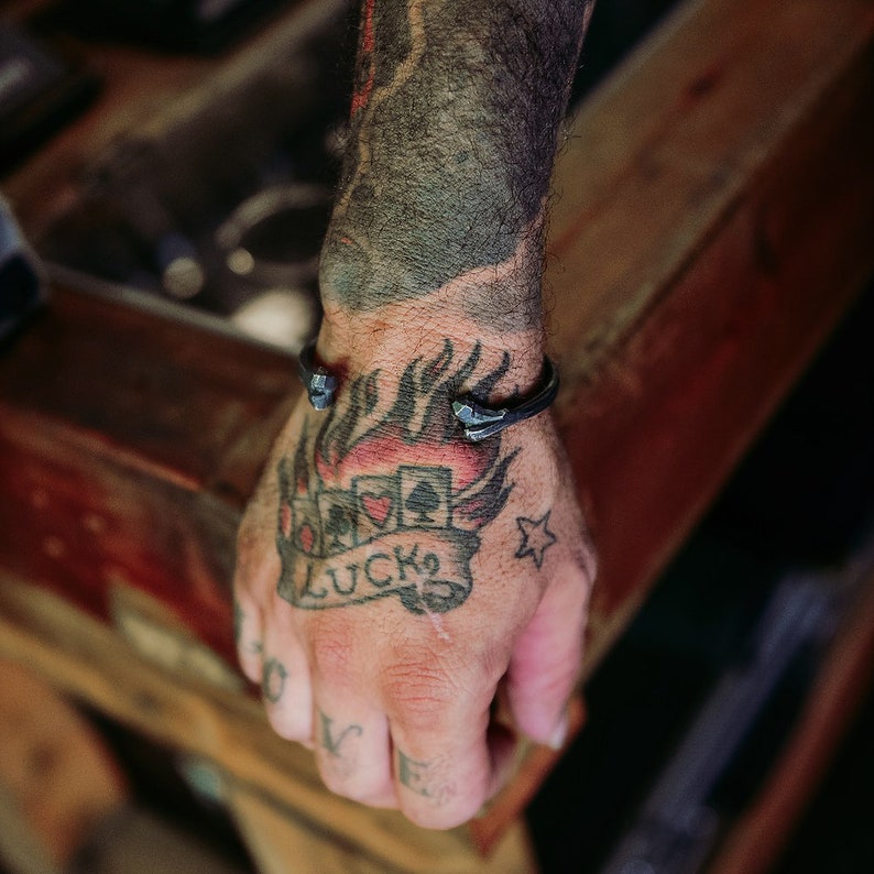 K&ouml;nnte beinhalten: Ein Arm mit einem Tattoo von Flammen, Spielkarten und dem Wort "LUCK" auf einem Banner. Die Person tr&auml;gt ein schwarzes Metallarmband mit Spikes.