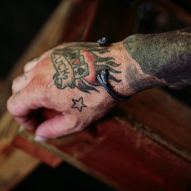 K&ouml;nnte beinhalten: Ein silbernes Metallarmband mit geh&auml;mmerter Oberfl&auml;che und einem spitzen Design. Das Armband wird an einem Handgelenk mit einem Tattoo von Spielkarten und einem Stern getragen.