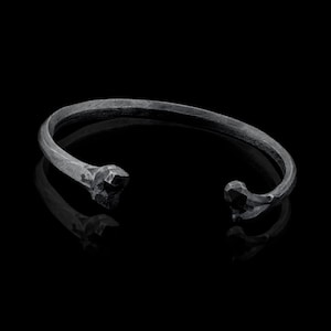 以下が含まれることがあります： 独特な角度のあるデザインの黒い金属製のブレスレット。ブレスレットは開いており、シンプルでミニマルなスタイルです。