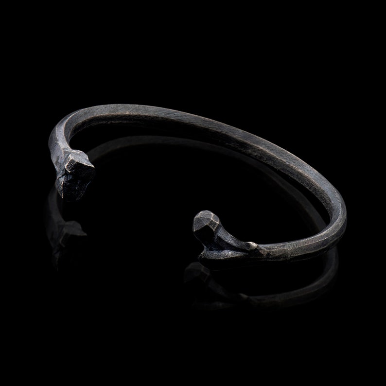 K&ouml;nnte beinhalten: Ein schwarzes Metallarmband mit einem einzigartigen, eckigen Design. Das Armband ist offen und hat einen schlichten, minimalistischen Stil.