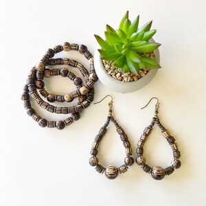 May include: A set of three brown wooden bead bracelets and a pair of brown wooden bead earrings. The earrings are teardrop shaped and have a gold hook.