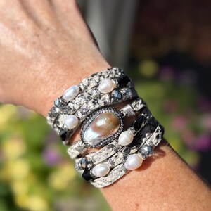 Peut inclure: Un bracelet multicouche avec un motif peau de serpent, orn&eacute; de perles blanches, de pierres noires et d'une grande pierre centrale ovale iris&eacute;e. Le bracelet est de couleur argent&eacute;e et s'enroule autour du poignet.