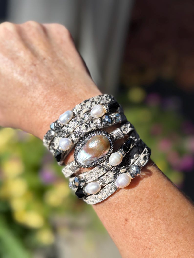 Peut inclure: Un bracelet multicouche avec un motif peau de serpent, orn&eacute; de perles blanches, de pierres noires et d'une grande pierre centrale ovale iris&eacute;e. Le bracelet est de couleur argent&eacute;e et s'enroule autour du poignet.