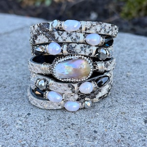 May include: Stacked bracelets with a snakeskin pattern, adorned with pearls and silver accents. The bracelets feature a mix of white, black, and silver tones, creating a layered look. The focal point is a large, iridescent pearl.