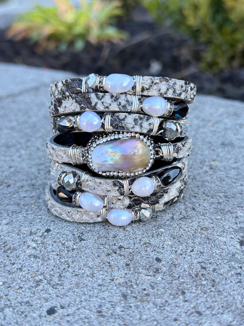 Peut inclure: Plusieurs bracelets superpos&eacute;s avec un motif peau de serpent, orn&eacute;s de perles et d'accents argent&eacute;s. Les bracelets pr&eacute;sentent un m&eacute;lange de tons blancs, noirs et argent&eacute;s. La pi&egrave;ce ma&icirc;tresse est une grande perle iris&eacute;e.