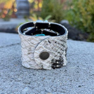 Peut inclure: Un bracelet manchette en cuir blanc et noir &agrave; motif peau de serpent avec une fermeture &agrave; bouton-pression argent&eacute;e. Le bracelet est rempli de divers bijoux argent&eacute;s et turquoise. Un accessoire unique.