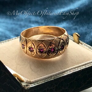 May include: A gold band ring with a textured design and red gemstones. The ring is in a small, black velvet box.