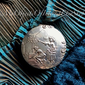 May include: A silver round compact with an embossed design of two figures in a garden setting. The figures are a woman holding a branch with leaves and a man kneeling on the ground. The compact is on a blue and green fabric background.