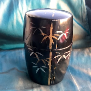 Op de afbeelding: Een zwarte gelakte cilindrische doos met een gouden en wit bamboe ontwerp. De doos is gesloten en heeft een deksel.