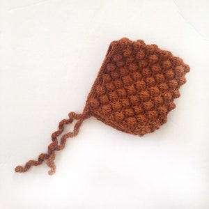 Peut inclure: Un bonnet marron crocheté avec un motif en point de boules texturé. Le bonnet a un bord festonné et de longues attaches torsadées. Le chapeau fait main est un accessoire chaud et confortable.