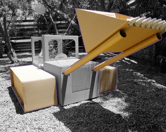 Puede incluir: Un juguete de bulldozer de cartón con una pala amarilla y un niño sentado en el asiento del conductor. El bulldozer está hecho de cartón corrugado y tiene un diseño simple.