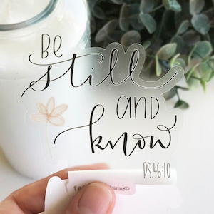 May include: A clear sticker with the text "Be still and know" in black cursive lettering with a small flower illustration. The text "PS. 46:10" is in smaller gray lettering below the main text.