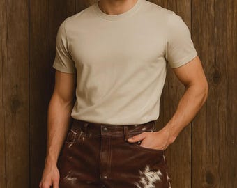 May include: A person wearing a beige crew neck t-shirt and brown leather shorts with white cowhide patches. The shorts have a brown leather belt and pockets. The background is a wooden wall.