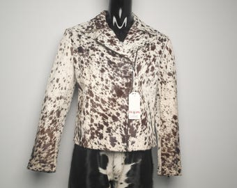 May include: A cropped jacket with a cowhide pattern in shades of brown and cream. The jacket features a notched collar, a diagonal zipper closure, and long sleeves. A tag hangs from the zipper. The jacket is displayed on a mannequin.