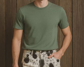 May include: A person wearing a short-sleeved, olive green t-shirt and denim shorts with a black and white spotted pattern. The background is a wooden wall.