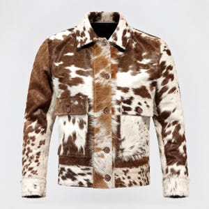 May include: A long-sleeve jacket featuring a brown and white cowhide print pattern. The garment includes a standard collar, a front button-down closure, and two chest pockets with buttoned flaps. The texture appears to be short-haired fur.