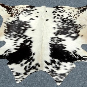 May include: A cowhide rug with a distinctive pattern of black spots and patches on a white background. The rug has an irregular shape with curved edges, adding a natural and rustic aesthetic. The hide is likely tanned and ready for use as a floor covering.
