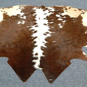Puede incluir: Una alfombra de piel de vaca con un patrón natural de marcas marrones y blancas. La alfombra tiene una forma única con bordes irregulares y un aspecto suave y texturizado. La piel es un revestimiento decorativo para el suelo.