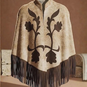 May include: A beige poncho with a textured, fur-like appearance featuring a dark brown floral applique design on the front. The garment has a high collar and a pointed hemline finished with long, dark brown fringe trim along the bottom edge.