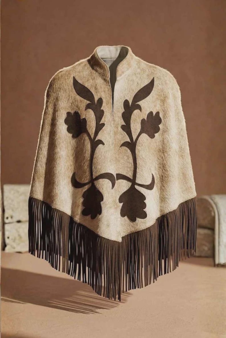 May include: A beige poncho with a textured, fur-like appearance featuring a dark brown floral applique design on the front. The garment has a high collar and a pointed hemline finished with long, dark brown fringe trim along the bottom edge.