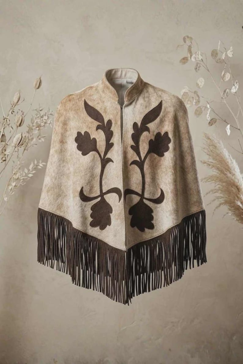 May include: A beige and tan patterned poncho featuring a high collar and a front zip closure. The garment is decorated with dark brown floral-shaped appliqu&eacute;s on the front panels and is finished with long dark brown fringe along the hem.