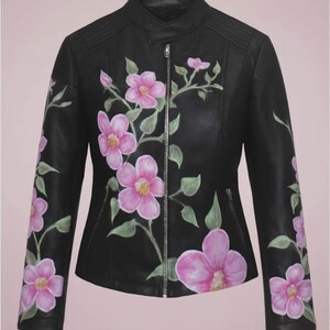 May include: Black leather jacket with a stand-up collar and a silver zipper. The jacket is adorned with hand-painted pink flowers and green leaves, creating a floral design. The sleeves and front of the jacket feature the floral pattern.