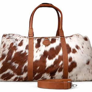 May include: A large duffel bag with a cowhide pattern in brown and white. The bag has brown leather handles, straps, and a detachable shoulder strap. It features side pockets and a buckle closure.