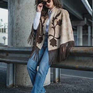 May include: A beige cape with dark brown floral patterns and long fringe trim, worn over a white button-down shirt, blue denim jeans, and dark brown heels. The outfit is accessorised with dark-tinted sunglasses.