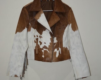 May include: A brown, white, and tan cowhide jacket with a zip-up front and fringe detailing on the sleeves. The jacket is hanging on a metal hanger. The jacket has a collar and a brown interior lining.