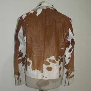 Pode incluir: Casaco de pele de vaca castanho e branco com gola cl&aacute;ssica e detalhes de z&iacute;per nas mangas. O casaco &eacute; exibido em um manequim, mostrando seu padr&atilde;o e textura &uacute;nicos. Uma pe&ccedil;a de roupa exterior elegante.