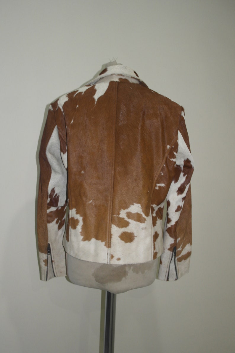 Pode incluir: Casaco de pele de vaca castanho e branco com gola cl&aacute;ssica e detalhes de z&iacute;per nas mangas. O casaco &eacute; exibido em um manequim, mostrando seu padr&atilde;o e textura &uacute;nicos. Uma pe&ccedil;a de roupa exterior elegante.