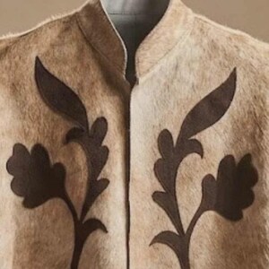 May include: A beige waistcoat with a textured, fur-like appearance featuring a high collar and dark brown, leaf-shaped appliqu&eacute; designs on both sides of the front opening. The garment has a light grey interior lining visible at the neckline.