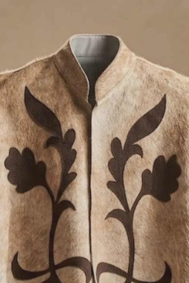 May include: A beige waistcoat with a textured, fur-like appearance featuring a high collar and dark brown, leaf-shaped appliqu&eacute; designs on both sides of the front opening. The garment has a light grey interior lining visible at the neckline.
