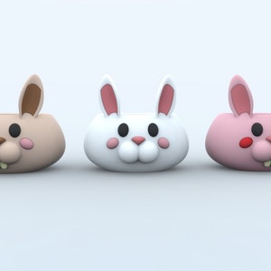 Könnte beinhalten: Drei Keramik-Blumentöpfe in Form von Hasen in verschiedenen Farben: Braun, Weiß und Rosa. Jeder Topf hat ein süßes Cartoon-Hasengesicht mit schwarzen Augen, einer braunen Nase und einem rosa Mund.
