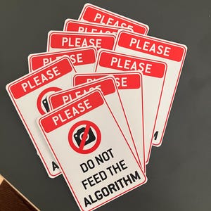 Può includere: Un gruppo di adesivi rettangolari bianchi con bordi rossi. Ogni adesivo presenta la parola "PLEASE" in alto e la frase "DO NOT FEED THE ALGORITHM" in basso, insieme a un simbolo di divieto di fotocamera.
