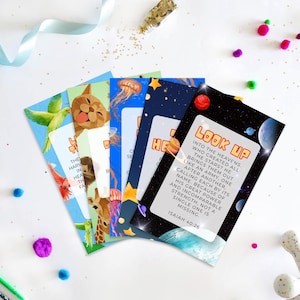 May include: A set of five rectangular cards featuring colourful illustrations of a green parrot, a cat, a giraffe, jellyfish, and a space scene. The top card displays the text LOOK UP INTO THE HEAVENS, WHO CREATED ALL THE STARS? ISAIAH 40:26.